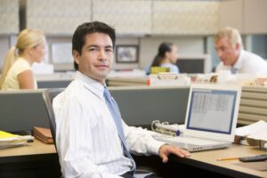 A male worker sits at a cubicle surrounded by other people with a white laptop on his desk